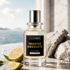 A bottle of Valued Scents 'Imagine Absolute' Eau de Parfum stands on a marble surface with a blurred seaside background, flanked by fresh lemon slices and rough white rocks. The clear glass bottle contains a pale golden liquid and features a black label with gold lettering and a silver cap. inspired by louis vuitton imagination - Imagine Absolute by Vaulted Scents - Designer inspired luxury fragrances, perfumes, aftershaves made in uk